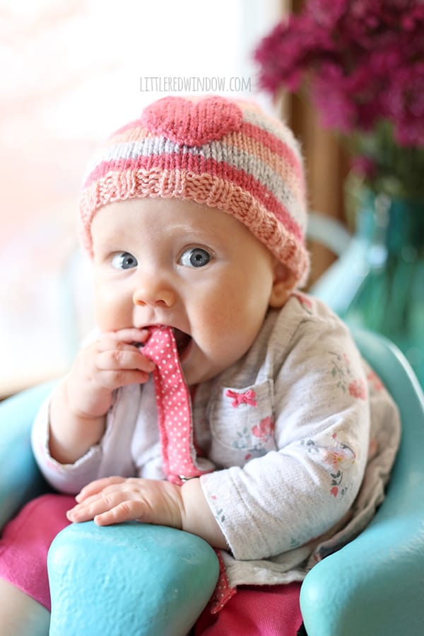 Chubby baby wearing pink striped hat with a heart on the front sitting in a seat and chewing the hem of her sweatshirt
