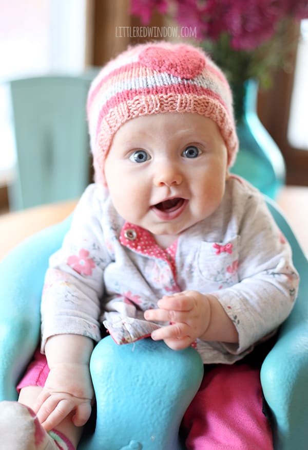 Chubby baby with mouth open wearing pink striped hat with a heart on the front sitting in a seat