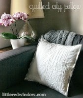 Quilted City Pillow sitting on a couch next to lamp and pot of pink flowers
