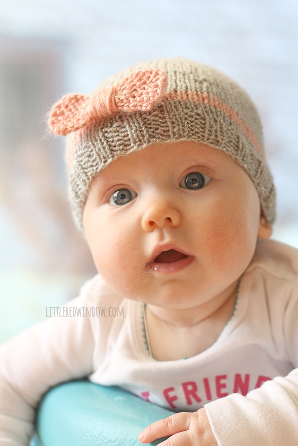 chubby baby with mouth open and wide blue eyes wearing gray knit hat with pink bow on the front