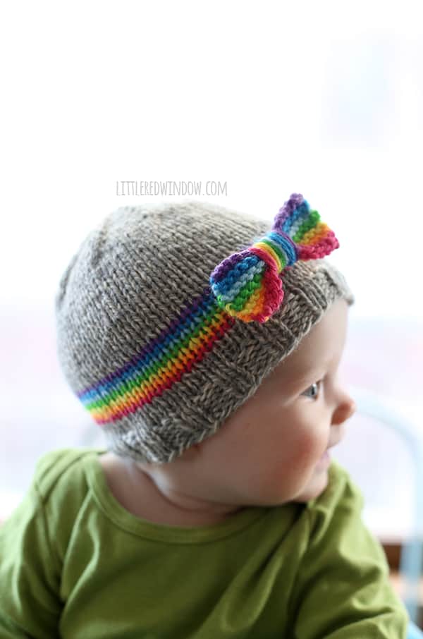 chubby baby in green shirt looking off to the right and wearing gray knit hat with rainbow stripe around the middle and rainbow bow on the front