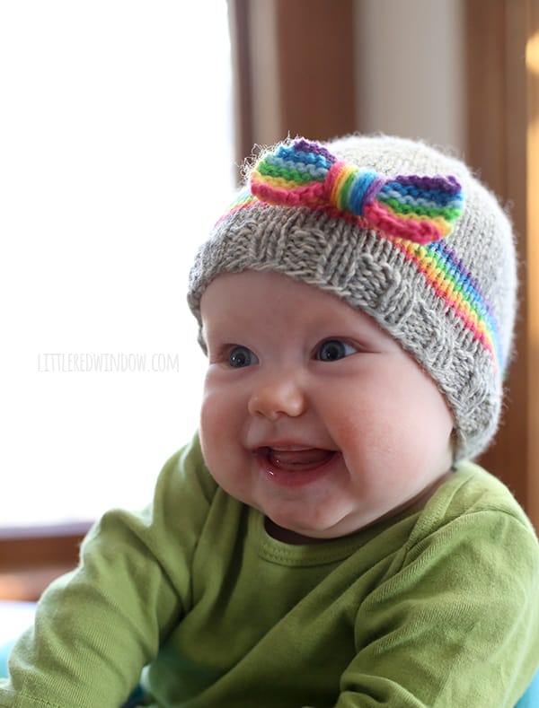 Smiling toothless baby in green shirt wearing gray knit hat with rainbow stripe and rainbow bow