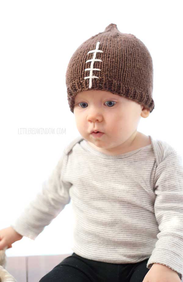 baby in gray shirt and brown knit hat that looks like a football looking down
