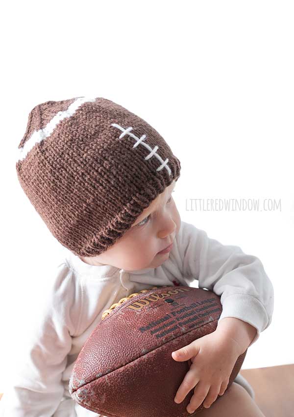 baby wearing brown football hat leaning forward and holding a real football under her arm