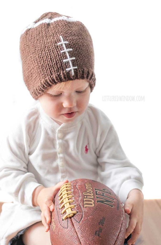 baby wearing brown knit football hat looking down at a real football in her lap