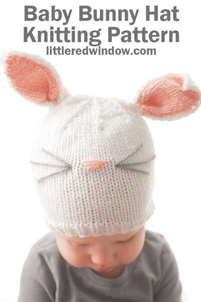 baby in gray shirt wearing a white knit hat with white and pink bunny ears on top and a pink nose and whiskers on the front looking down at their lap