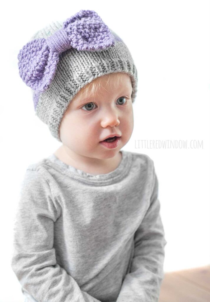 child wearing a gray shirt and gray knit hat with a purple band around the middle a large purple seed stitch bow on the front looking off in the distance to the near right