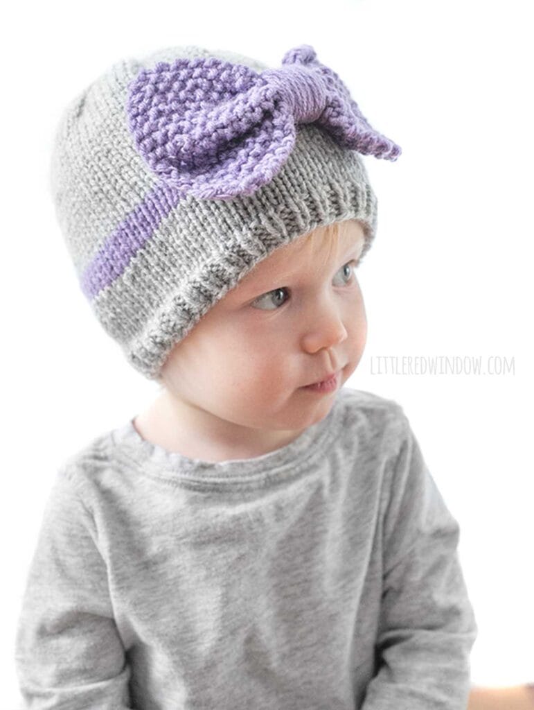 child wearing a gray shirt and gray knit hat with a purple band around the middle a large purple seed stitch bow on the front looking slightly to the right