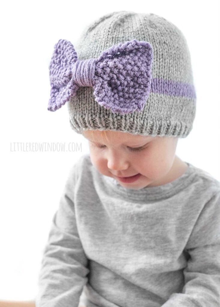 child wearing a gray shirt and gray knit hat with a purple band around the middle a large purple seed stitch bow on the front smirking and looking down at their hands