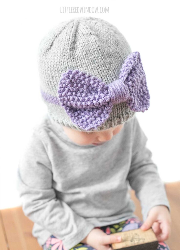 view from slightly above of child wearing a gray shirt and gray knit hat with a purple band around the middle a large purple seed stitch bow on the front smirking and looking down at their hands