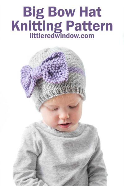 child wearing a gray shirt and gray knit hat with a purple band around the middle a large purple seed stitch bow on the front looking down at their lap in front of a white background