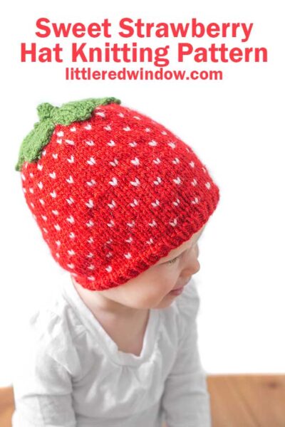 toddler in white shirt wearing a red knit strawberry hat with white strawberry seed polka dots and green leaves on top looking off to the right