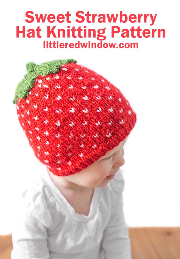 toddler in white shirt wearing a red knit strawberry hat with white strawberry seed polka dots and green leaves on top looking off to the right