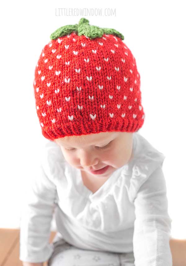 toddler in white shirt wearing a red knit strawberry hat with white strawberry seed polka dots and green leaves on top looking down and reaching forward