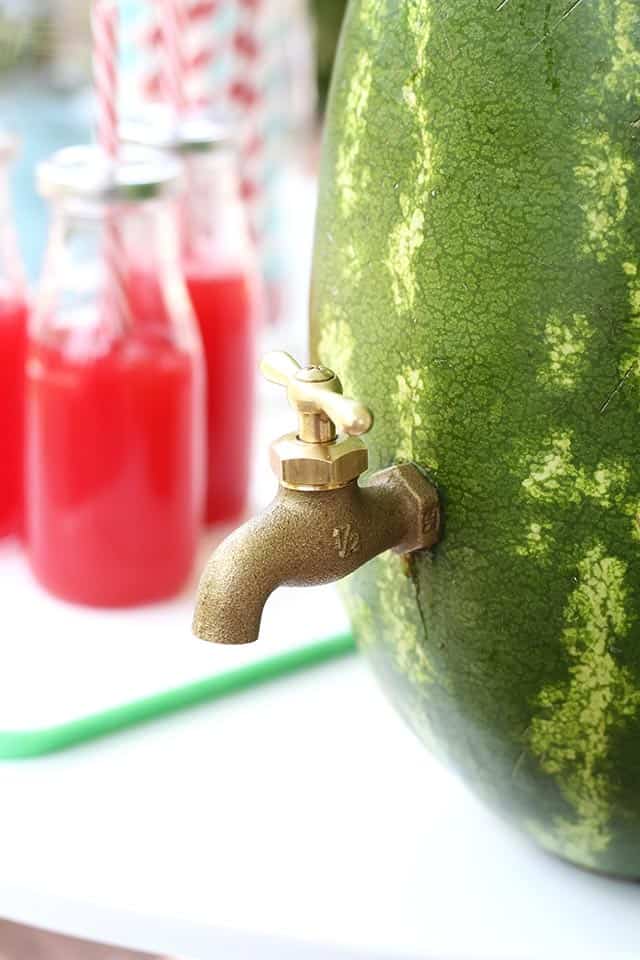 Closeup of a watermelon with a spigot in the side to be used as a drinks dispenser. Glasses of red juice in the backgournd