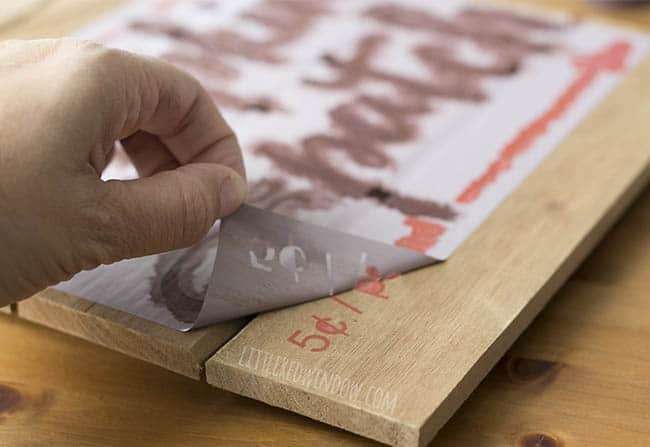 hand peeling off the stencil to show the painted design on the wood sign underneath