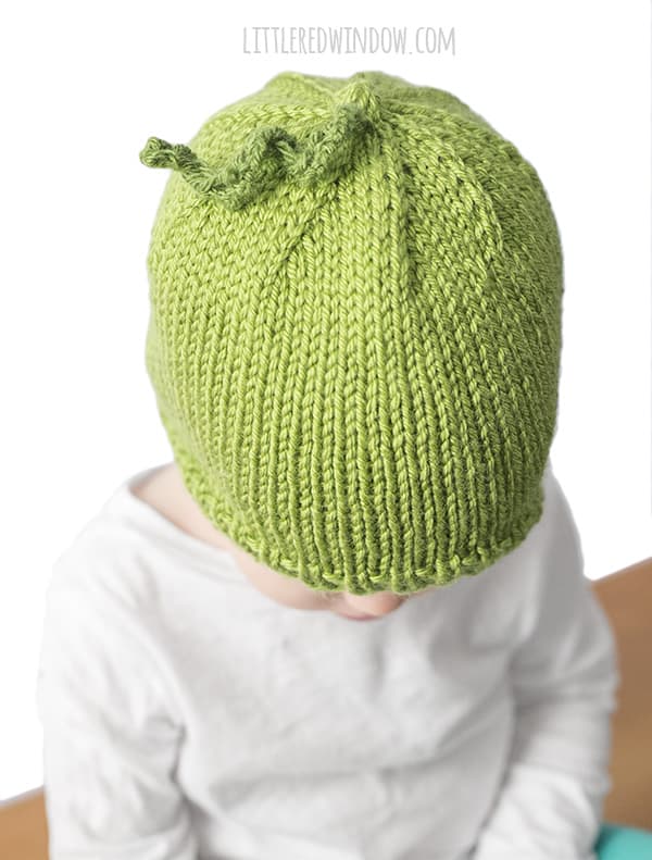 top view of little girl wearing a green pea shaped knit hat and a white shirt 