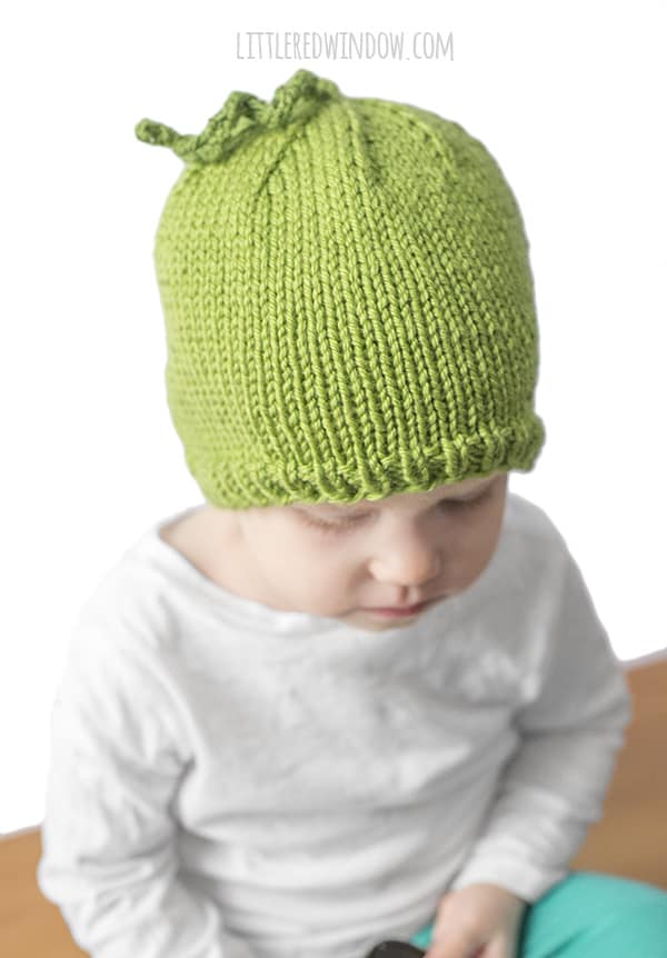 little girl wearing a green pea shaped knit hat and a white shirt and looking down