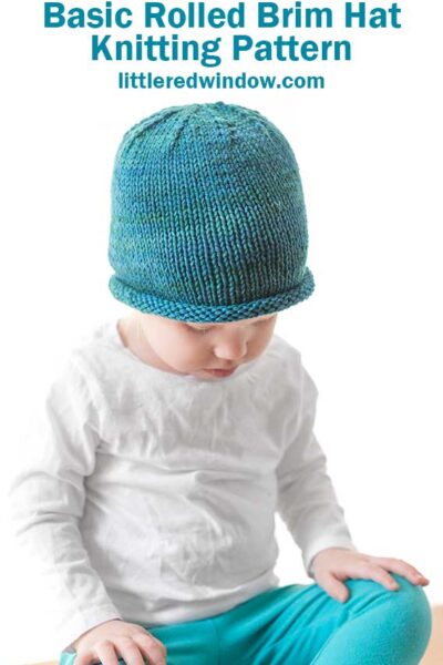 toddler in white shirt and blue pants wearing a knit hat with a rolled brim made of teal blue hand dyed yarn sitting on a wood table and looking down at their lap