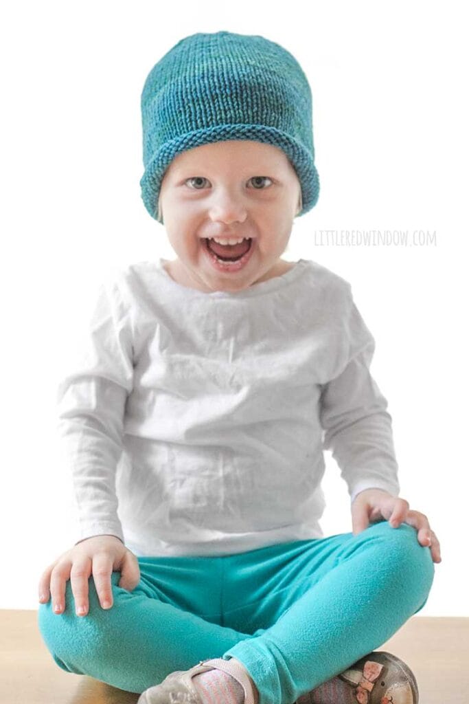 toddler in white shirt and blue pants wearing a knit hat with a rolled brim made of teal blue hand dyed yarn sitting on a wood table and smiling at the camera
