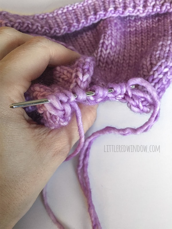 Hand holding the knit flat baby hat with the last 9 stitches threaded onto the yarn needle to finish