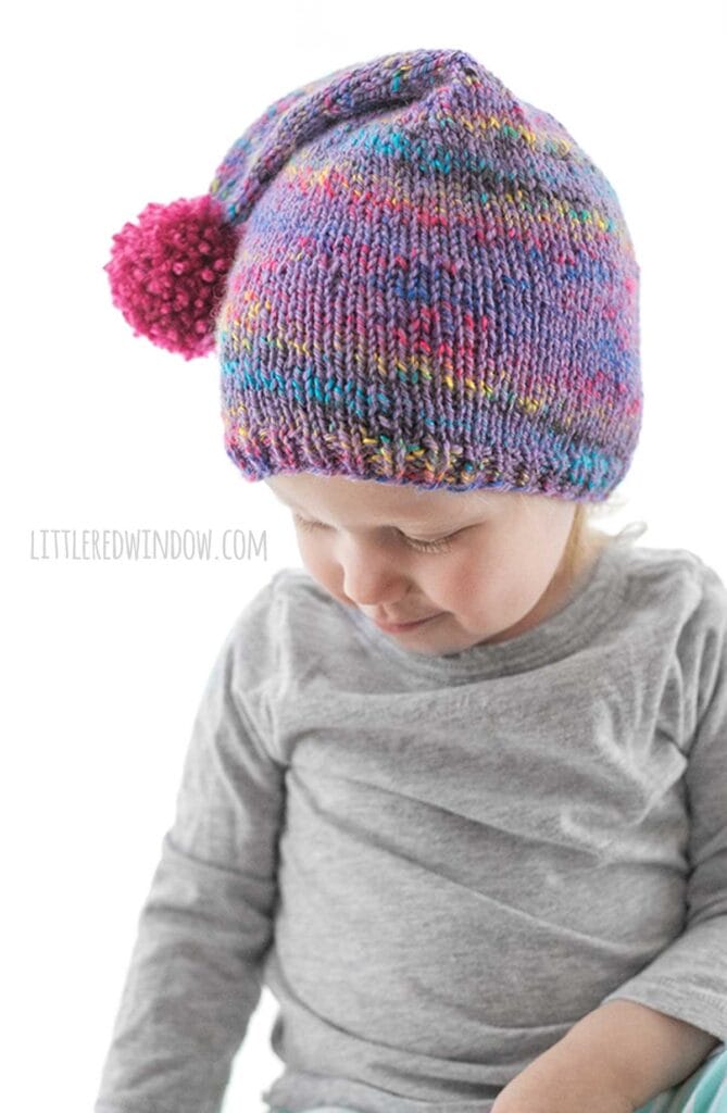 child in gray shirt with wearing a purple speckled knit stocking cap with magenta pom pom hanging to the side while they look down