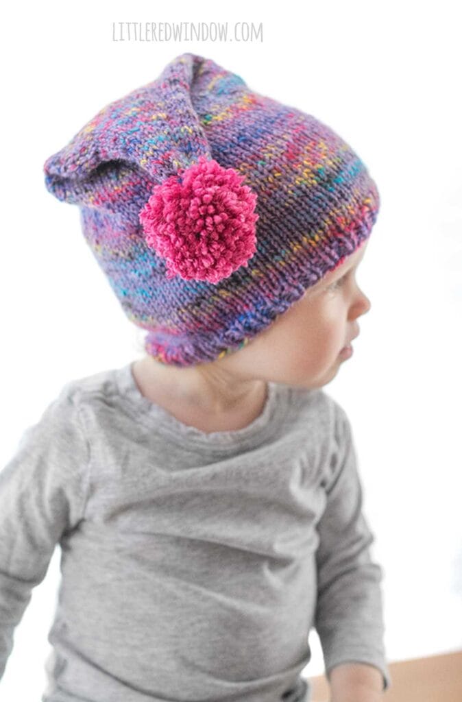 side view of child in gray shirt with wearing a purple speckled knit stocking cap with magenta pom pom hanging to down while they look to the right
