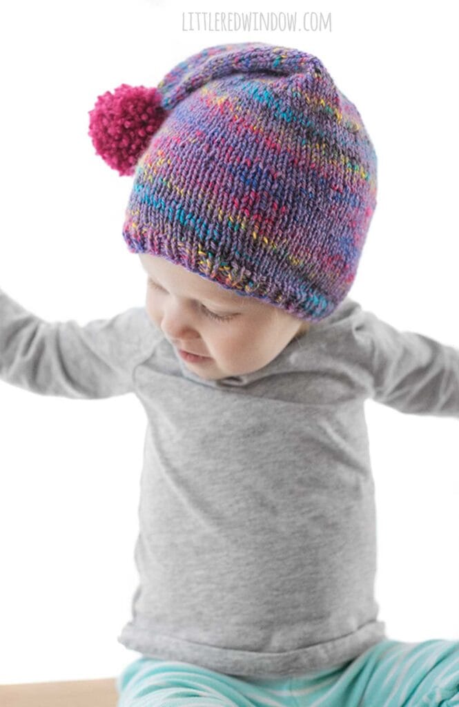 child in gray shirt with arms out to the side wearing a purple speckled knit stocking cap with magenta pom pom on it