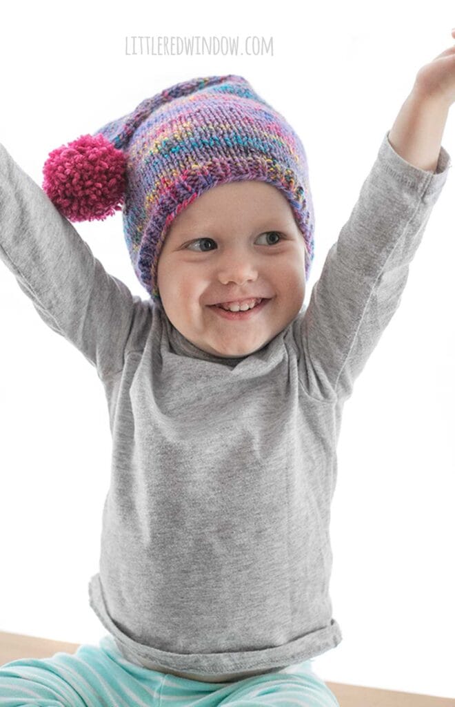 happy child with arms up in the air wearing a gray shirt and purple speckled knit stocking cap with hot pink pom pom