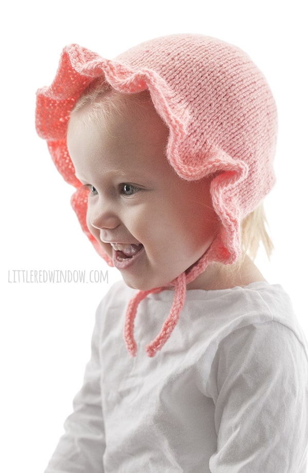 side view of laughing chubby toddler in white shirt in front of a white background wearing a light pink ruffle bonnet knitting pattern with chin ties and looking off to the left