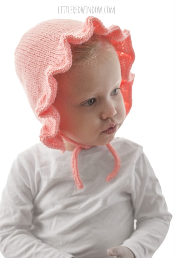 front view of chubby toddler in white shirt in front of a white background wearing a light pink ruffle bonnet knitting pattern with chin ties and looking slightly off to the right while making a kissy face