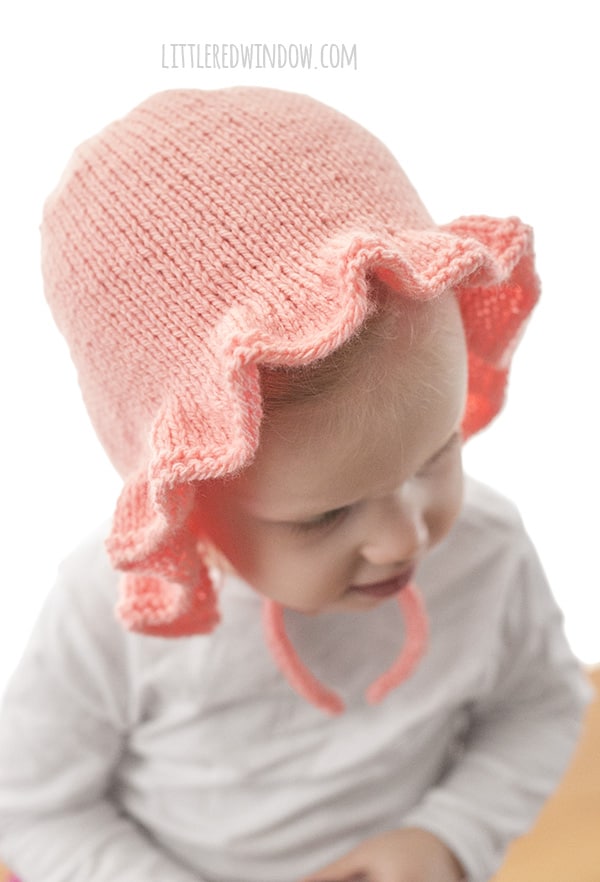 view from slightly above of chubby toddler in white shirt in front of a white background wearing a light pink ruffle bonnet knitting pattern with chin ties and looking down to show the ruffles clearly