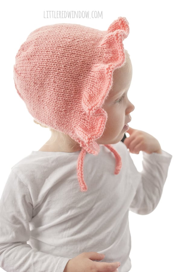 side view of chubby toddler in white shirt in front of a white background wearing a light pink ruffle bonnet knitting pattern with chin ties and looking off to the right with one arm up pointing at their face