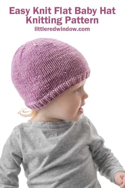 toddler in gray shirt wearing a lavender knit hat in front of a white background looking far off to the right