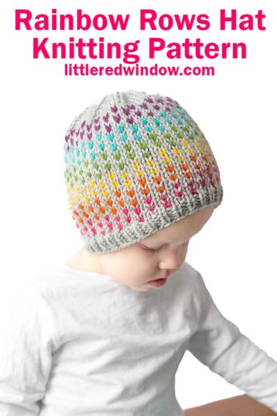 A child wears a knitted hat adorned with rainbow colors and a gray brim, perfectly capturing the charm of the "Rainbow Dots Hat Knitting Pattern" from littleredwindow.com. With a white shirt on, the child gazes downward, enhancing the endearing scene.