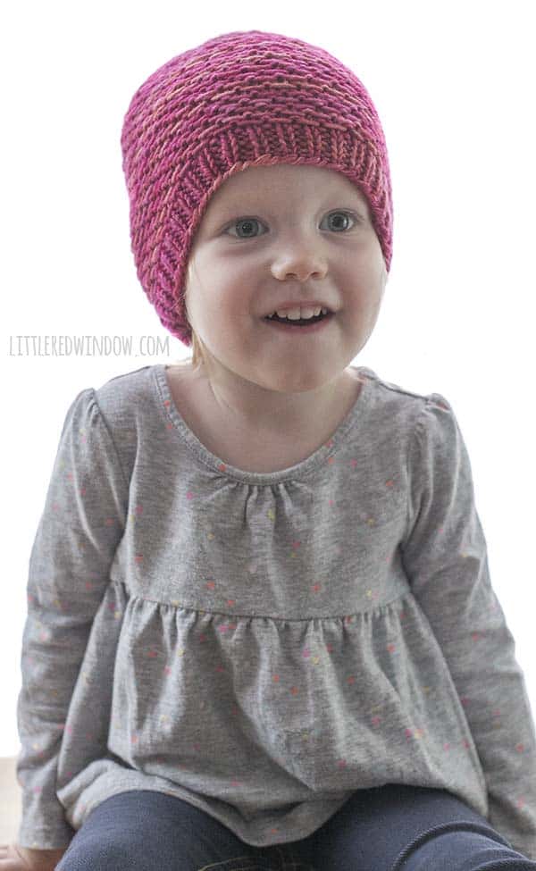 Little girl in gray shirt smiling and wearing magenta colored knit slipped purl stitch hat