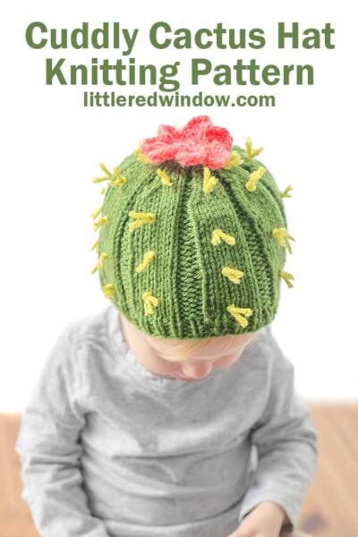 Child in gray shirt on a wood table wearing a medium green ribbed knit hat with light green yarn spikes and a pink cactus flower on top to make a cactus hat looking down at their lap
