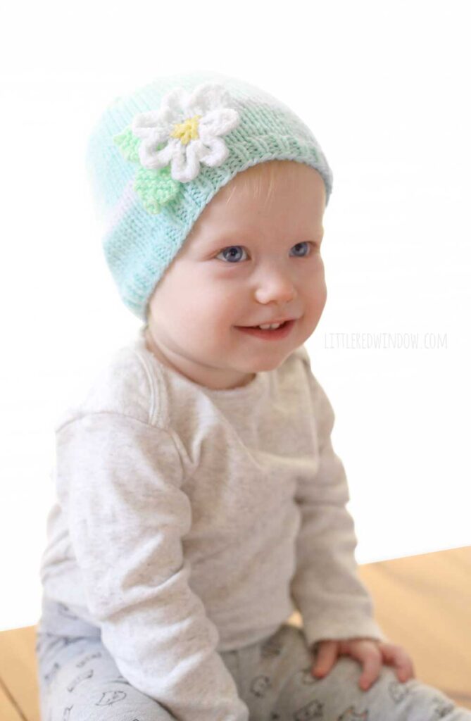 Baby in gray shirt smiling and wearing a light blue spring daisy flower hat with white band and white knit flower in front of a white background