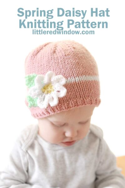 baby in gray onesie examining something in their hands while wering a light pink knit hat with a white stripe around the middle and white knit flower on the front of the hat in front of a white background