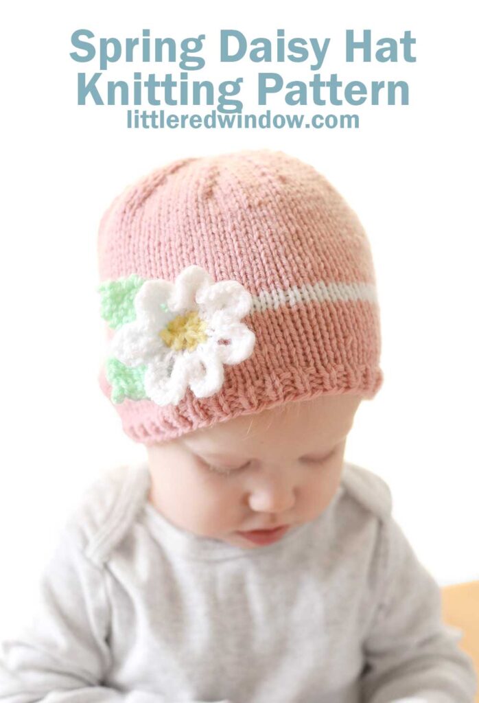 baby in gray onesie examining something in their hands while wering a light pink knit hat with a white stripe around the middle and white knit flower on the front of the hat in front of a white background