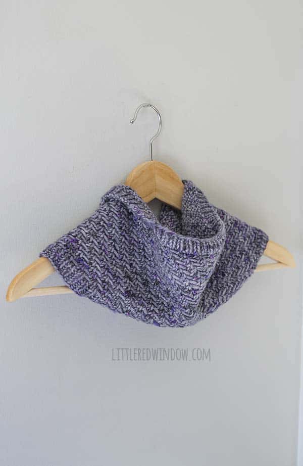 purple knit cowl hanging on a light wood hanger in front of a light gray wall