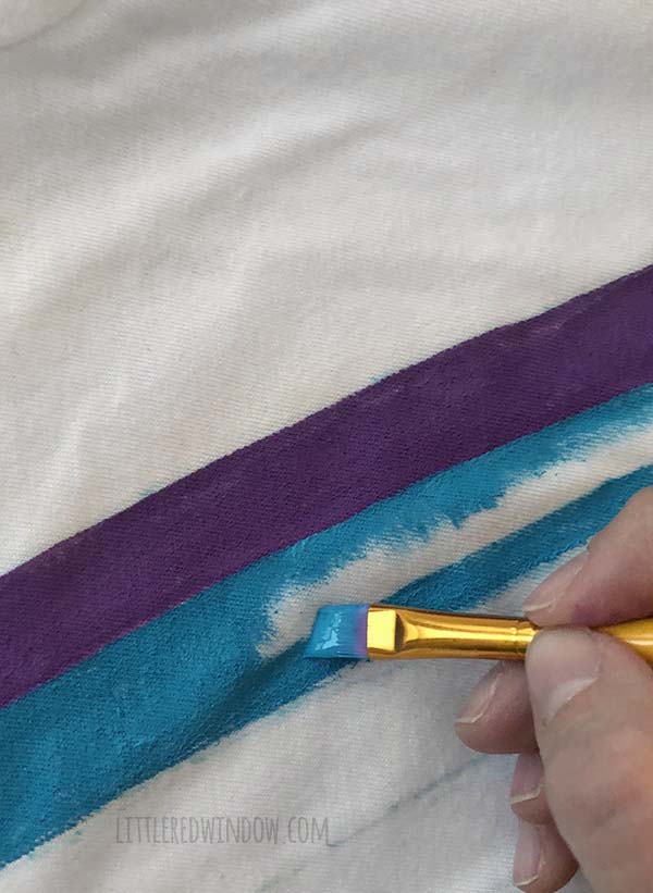 A hand painting a blue stripe of the rainbow on a white tshirt
