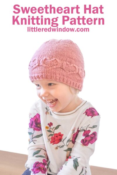 A smiling child dons a pink knitted "Sweetheart Hat" paired with a floral-patterned shirt. Text above reads: "Sweetheart Hat Knitting Pattern" with a website link below.