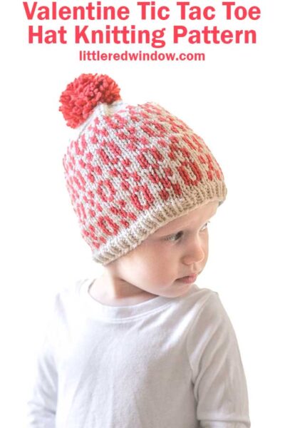 A child wears a knitted Tic Tac Toe Hat adorned with red and white xo patterns and a red pom-pom. Above, the text reads "Valentine Tic Tac Toe Hat Knitting Pattern" in vibrant red. The child complements the look with a crisp white shirt.