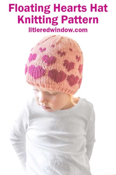 A child in a pink knitted hat adorned with heart patterns gazes downward. The text at the top reads, "Floating Hearts Hat Knitting Pattern litleredwindow.com." Dressed in a white shirt against a simple backdrop, they showcase the charm of this adorable hearts hat knitting pattern.
