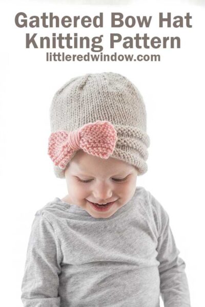 smiling girl in gray shirt wearing tan knit hat gathered in the front with large pink knit bow looking down at her lap