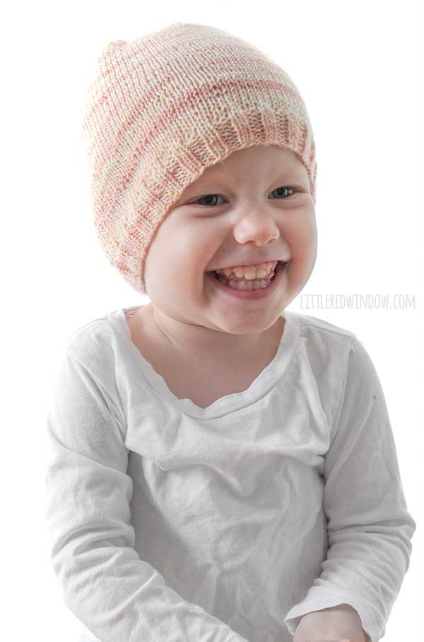 laughing baby in white shirt wearing a light pink lightly striped sock weight yarn knit hat