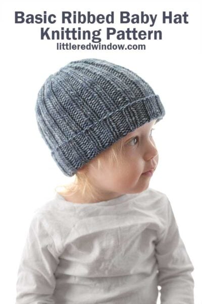 small child in gray shirt wearing a medium denim blue colored ribbed knit hat with folded up brim looking off to the right in front of a white background
