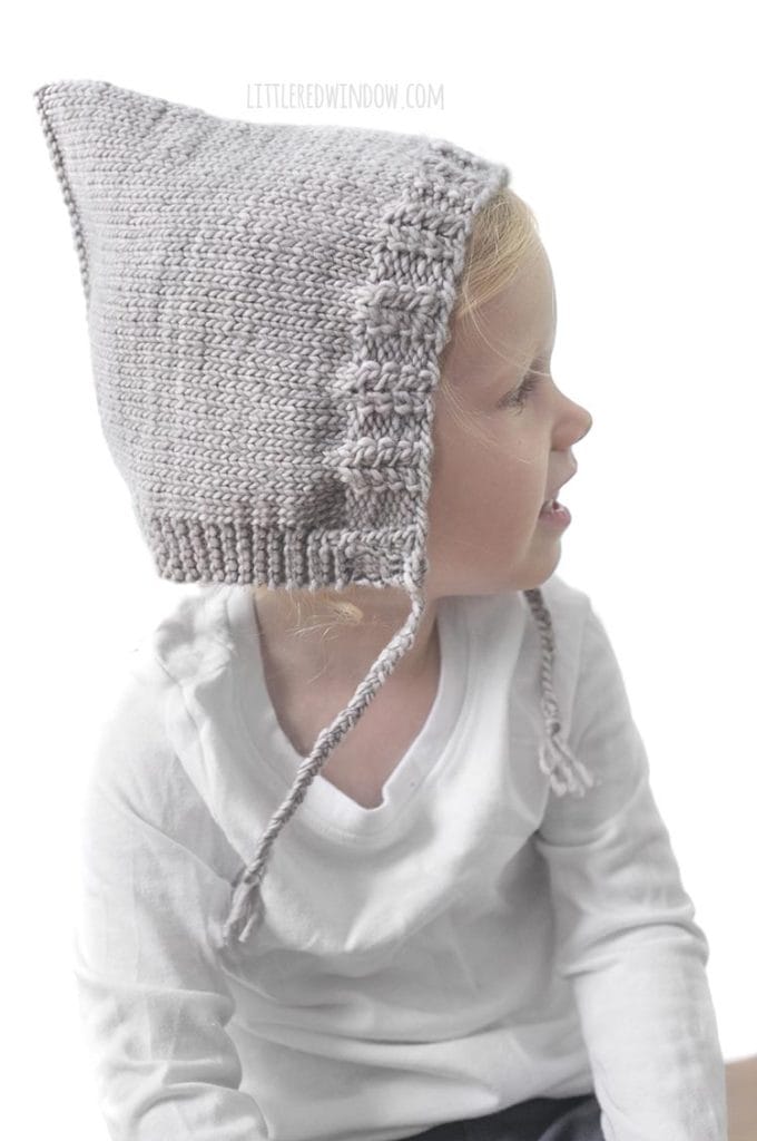 side view of little girl in white shirt wearing tan knit pixie bonnet with braided chin ties looking to the right