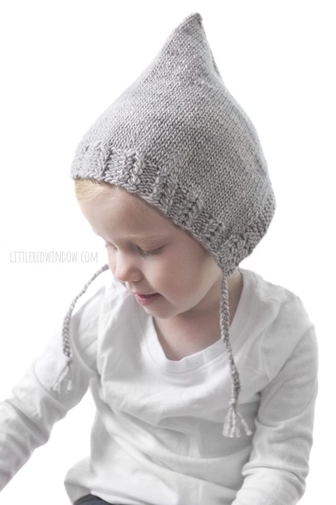 little girl in white shirt wearing tan knit pixie bonnet with braided chin ties looking down and to the left